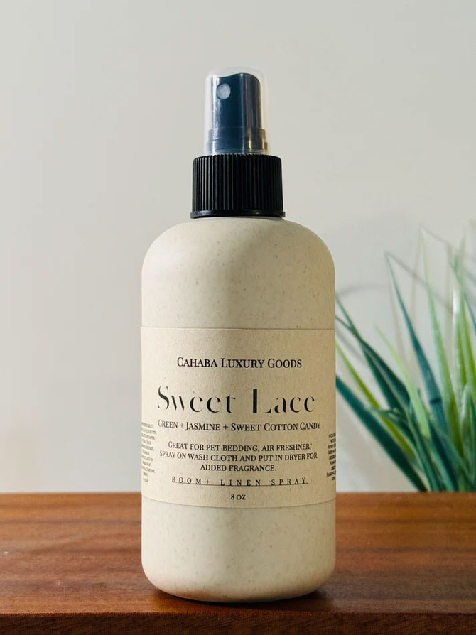 Cahaba Luxury Goods Room Spray, an 8 oz bottle with a spray nozzle containing a sweet lace scent with green jasmine and sweet cotton candy, on a wooden surface.