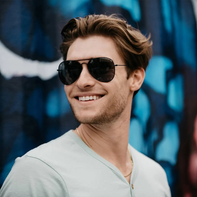 Knockaround Mile Highs SM Sunglasses, stylish black sunglasses worn by a smiling young man with a graffiti background.