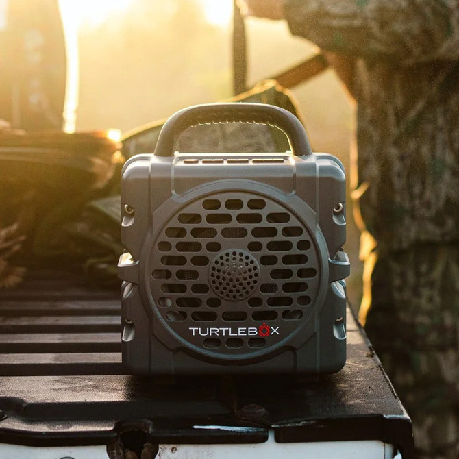 Turtlebox Audio Turtlebox Cub, portable outdoor speaker with rugged design and grille front, on a wooden surface during sunset.