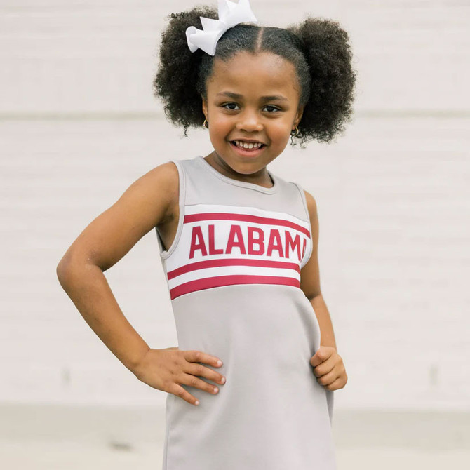 Scarlet and Gold Youth The Classic Cheer Dress Alabama, a young girl wearing a gray cheerleading dress with red ALABAM logo and stripes, smiling with hands on her hips and white bow in her curly hair.