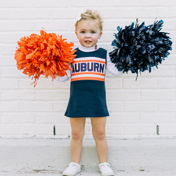 Scarlet and Gold Youth The Classic Cheer Dress Auburn, a young girl wearing a navy blue cheerleading dress with Auburn logo and white sleeves, holding orange and navy blue pom-poms against a white brick wall.