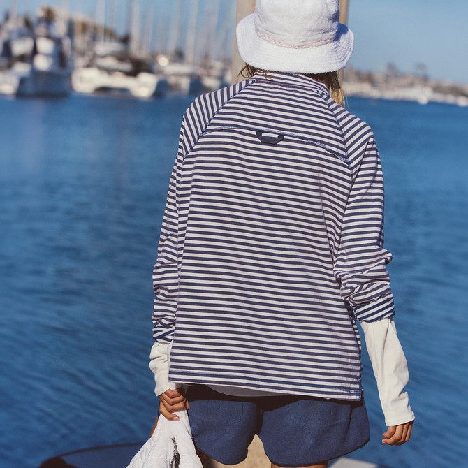 FP Movement Women's Restoration Yarn Dye Stripe Layer, image of a woman wearing a navy and white striped layer with a white hat near the water.
