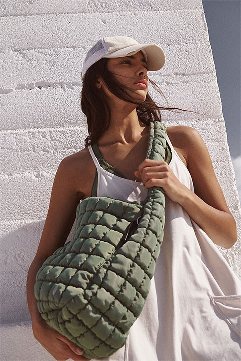 FP Movement Quilted Carryall, woman with long brown hair wearing a white cap and tank top, holding a large quilted green carryall bag on her shoulder against a white brick wall.