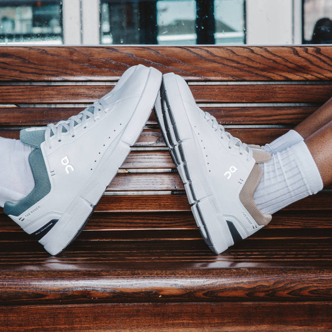 On Running Men's The Roger Advantage, white athletic sneakers with contrasting heel colors, placed on a wooden bench with a city background.