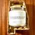 Cahaba Luxury Goods Cahaba Luxury Candles, white candle with gold lid in a transparent jar with a label, in a woven basket on a wooden surface.