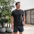 Rhoback Men's Blitz Tech Tee, a man wearing a black athletic T-shirt outdoors with plants and a modern building in the background.