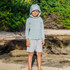 Free Fly Apparel Free Fly Toddler Breeze Short, a young boy wearing a light gray long sleeve hooded shirt and matching shorts standing on a sandy and rocky beach with green foliage in the background.
