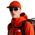 The North Face Runner Trucker, a man wearing an orange mesh-back trucker cap with a flat brim, white sporty sunglasses, and an orange waterproof jacket with a backpack, highlighting outdoor adventure gear.