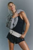FP Movement Women's Carpe Diem Runsie, a woman wearing a black athletic tank top, black shorts with a white waist bag, and a gray hoodie draped over her shoulders.