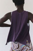 FP Movement Women's Tempo Tank, back view of a woman wearing a purple sleeveless athletic tank top with a white sports bra underneath, paired with colorful patterned workout shorts.