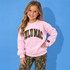 Lulu Mac Youth Mossy Oak Embroidered Applique Crew Sweatshirt, model wearing a pink crewneck sweatshirt with Mossy Oak camouflage Lulu Mac lettering and a sleeve label, paired with camouflage pants against a blue background.