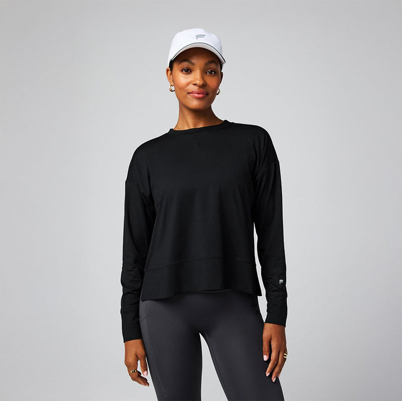 Fabletics Women's Cloud Jersey Boxy Long-Sleeve, a woman wearing a loose black long-sleeve athletic top with a relaxed fit and high-low hem, paired with dark leggings and a white cap, standing against a plain light gray background.