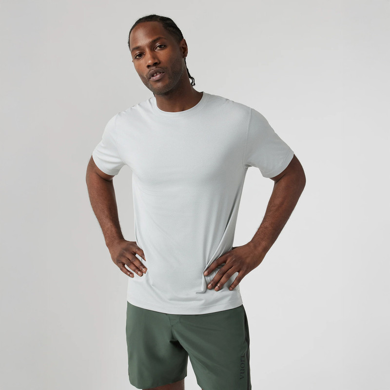 Vuori Men's Tradewind Performance Tee 2.0, a man wearing a white athletic t-shirt and green shorts posing with hands on hips against a plain background.