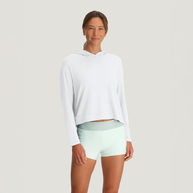 Free Fly Women's Bamboo Shade Crossover Hoodie, a woman in a white hoodie with a crossover design and long sleeves, paired with pastel green shorts, standing against a plain light background.