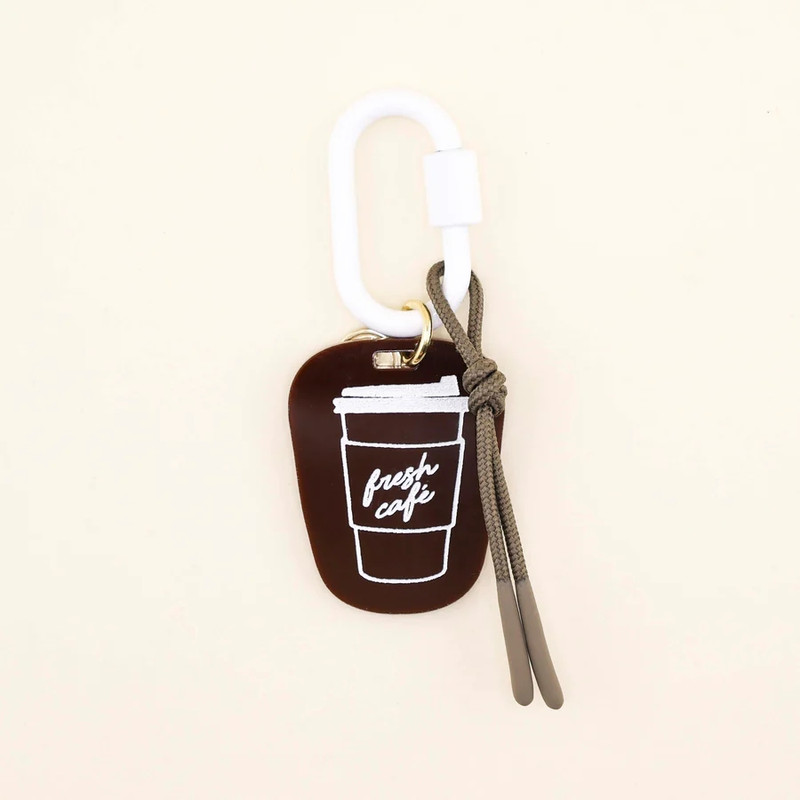 Freshwater Curated Goods Coffee Bag Charm, a brown silicone coffee cup with white line drawing of a coffee cup and the text 'fresh cafe' on it, attached to a white carabiner and gray shoelace pendant.