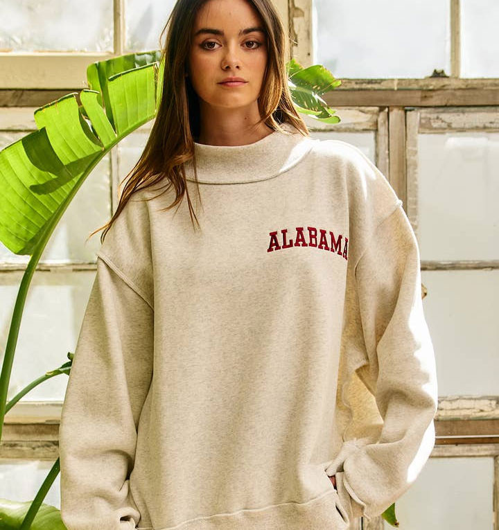 Bucketlist Women's Alabama Embroidered Mock Neck Sweatshirt, beige sweatshirt with red 'Alabama' embroidery, casual style, worn by a woman in a bright indoor setting with greenery.