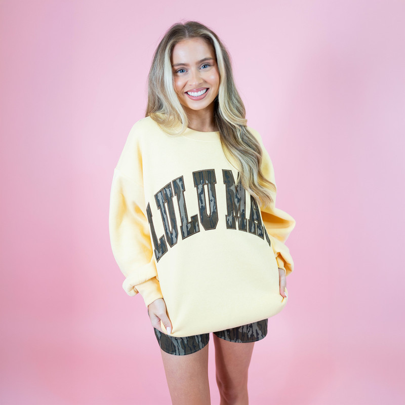 Lulu Mac Women's Mossy Oak Embroidered Applique Crew Sweatshirt, model wearing a pale yellow crewneck sweatshirt with large Mossy Oak camouflage lettering and camo shorts against a pink background.