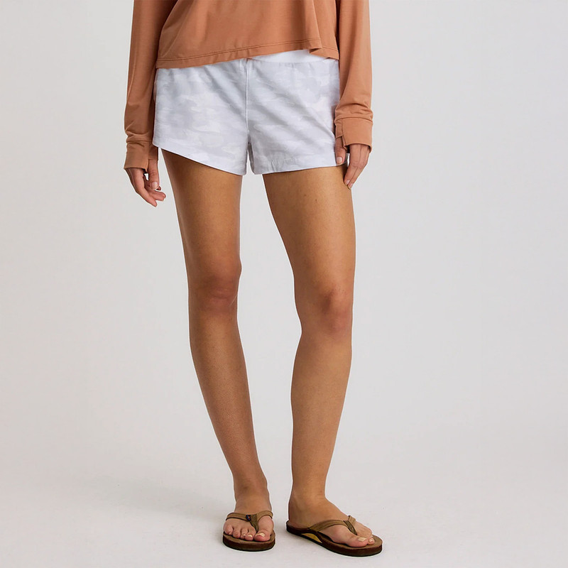 Free Fly Apparel Women's Bamboo-Lined Active Breeze Short - 3" Inseam, white shorts with camouflage pattern and bamboo lining, worn by a woman with a tan shirt and flip flops.