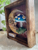 Angled view of Charming highlighting the dimensional clay moon face, whimsical mushroom, handcrafted details, and depth of the wooden frame.