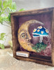 Angled view of Charming highlighting the dimensional clay moon face, whimsical mushroom, handcrafted details, and depth of the wooden frame.