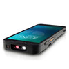 Low-angle perspective view of a black Datalogic Memor 30 mobile computer resting flat on a reflective white surface, showcasing the active top scanning window. The illuminated scanner aperture reveals bright white LEDs and an active red laser crosshair or barcode aiming pattern. To the left of the scanning lens is a rubberized power button with an orange power symbol. The device's right profile is visible, highlighting a textured volume rocker and a scan trigger button with a prominent yellow stripe. The large glass touch display is active with a blue-green lock screen layout reflecting subtly on the casing.