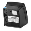 The file bixolon-srp-350plusiii-pos-thermal-receipt-printer-srp-350plusiiicosg-rdu-bottom.jpg displays the underside and rear-side profile of a black BIXOLON thermal receipt printer, which is resting tilted on its side against a white background.

The bottom panel features a large white manufacturer label with technical specifications, barcodes, and regulatory symbols, positioned upside down relative to the photo's orientation. Four circular rubber feet are placed at the corners of the base. A small rectangular silver metal access plate secured by a screw sits near the center, and a recessed RJ12 cash drawer port is located on the left side of the base. A blue Energy Star sticker is visible on the upper left corner of the housing, and a small black power switch is recessed near the back edge.