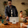 A rear-view product shot of a Square Stand for iPad (2nd generation) setup on a wooden counter, showing its white back panel and white base with the black Square logo. A male customer wearing glasses and a dark jacket over a floral shirt is swiping or dipping a payment card on the side of the terminal. In the background, a person pours coffee into a white mug, while another customer sits at a table against a gray brick wall with hanging plants.