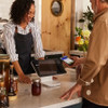 A smiling female business owner in a striped shirt and dark apron stands behind a marble counter, using a black Square Stand for iPad with a white base. A male customer holds his smartphone near the contactless card reader on the side of the stand to complete a mobile payment transaction. The register screen shows a total of $17.25. Glass jars, flowers, and a warm wooden wall are visible in the background.