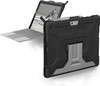 A product shot showcasing two views of the Microsoft Surface Go setup. In the foreground, the back of the device is featured, showing the black rugged texture and a silver, industrial-style built-in kickstand bearing the UAG logo. In the background, the tablet is propped up and connected to a grey type cover keyboard.