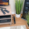 A Tripp Lite TLP606B surge protector placed on a hardwood floor next to a wooden media console and a large black tower speaker. Several power cords plug into the strip, which discreetly runs behind the home entertainment setup. Two remote controls rest on the console above.