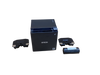 A matte black, cube-shaped Epson thermal receipt printer is centered on a plain, light brown cardboard surface. The front face features the "EPSON" logo in white text, a small wireless symbol near the top right, and an Energy Star sticker in the bottom right corner. On the top panel, two small buttons—a power button and a feed button—are visible on the right side next to a gray paper release lever. Flanking the printer are two identical, detached black plastic brackets or covers with silver metallic accents on their edges. A tightly wrapped, small roll of black thermal paper secured with white tape lies to the right of the printer.