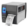 Three-quarter view of the printer in action, dispensing a printed shipping and barcode label through the front mechanism with a clear side window view of internal supplies.