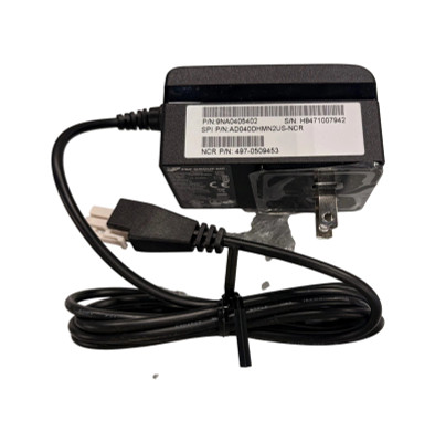 A full view of the black FSP Group AC power adapter against a white background. The power brick features a white label with barcodes and part numbers, including "NCR P/N: 497-0509453" and "SPI P/N: AD040DHMN2US-NCR." On the right side, a standard two-prong North American wall plug is visible, still covered in protective clear plastic. A black power cord extends from the left side of the brick, coiled and secured with a black cable tie, ending in a white 2-pin connector.