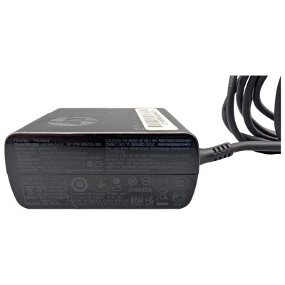 An eye-level shot of a rectangular, black Hewlett-Packard (HP) AC adapter. The adapter is positioned horizontally, showing its side profile with a detailed regulatory label and technical specifications. The top surface features a debossed HP logo and a small white barcode sticker. A thick, black power cord is attached to the right side of the adapter, curving upward and out of the frame. The background is a solid, clean white.