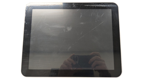 A front-facing, straight-on shot of a POSBANK APEXA 12.1-inch customer display. The monitor has a thick black bezel with rounded corners and a thin white outer casing. The screen is powered off and shows significant surface smudging, fingerprints, and a reflection of a person holding a smartphone to take the photo.