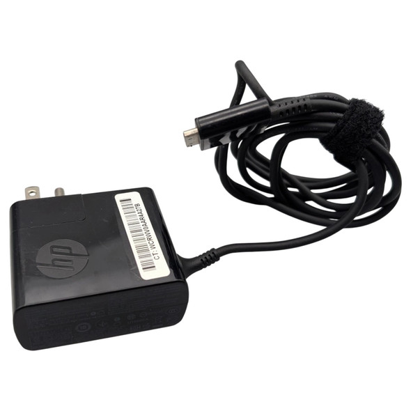 An overhead, slightly angled shot of a black HP AC adapter and its coiled power cable on a white background. The square adapter block features a debossed HP logo and a white barcode sticker on its top surface, with two metal power prongs visible at the top edge. A black cable extends from the side of the block, leading to a coiled bundle secured by a black Velcro strap. The cable ends in a proprietary, multi-pin HP tablet connector shown in the upper center of the frame.