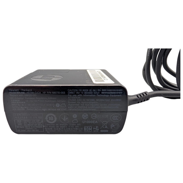 An eye-level shot of a rectangular, black Hewlett-Packard (HP) AC adapter. The adapter is positioned horizontally, showing its side profile with a detailed regulatory label and technical specifications. The top surface features a debossed HP logo and a small white barcode sticker. A thick, black power cord is attached to the right side of the adapter, curving upward and out of the frame. The background is a solid, clean white.
