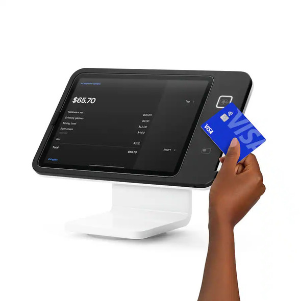 A product shot of a black Square Stand for iPad (2nd generation) on a white base, isolated against a white background. The terminal's dark checkout screen displays a transaction total of $65.70. A person's hand is shown dipping a blue Visa credit card into the chip reader slot on the right side of the frame, with an illuminated contactless payment icon visible just above it.