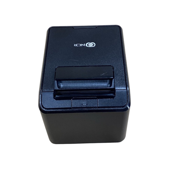Straight-down top view of a matte black cubical NCR RealPOS 7199 thermal receipt printer, highlighting the clean white NCR logo printed on the paper roll cover, the integrated plastic cover release latch, and a flat front profile, isolated on a white background.