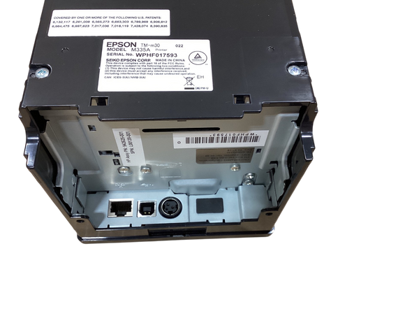 An inverted view of a black Epson TM-m30 thermal receipt printer, model M335A. The image displays the underside connection panel, which features an Ethernet port, a USB Type-B port, and a circular power connector, alongside the device's main compliance label, a serial number sticker, and additional manufacturing assembly information.