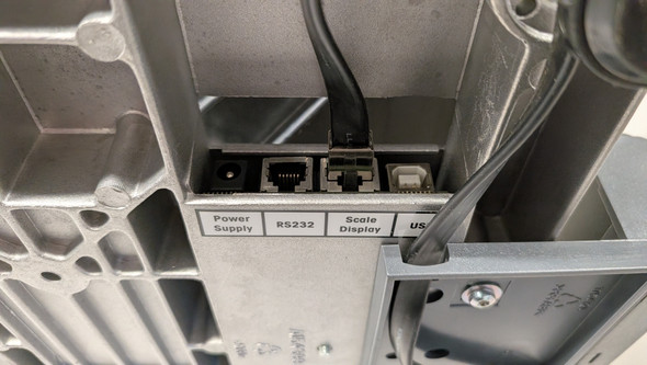 A close-up view of the ribbed cast-aluminum underside of a Mettler Toledo PS60 scale, highlighting the recessed port bank. A white labels grid marks the connection types from left to right: "Power Supply" (empty barrel jack port), "RS232" (empty RJ port), "Scale Display" (occupied by a flat black modular communication cable running down from the scale base), and a square "USB" port. A thick black cable wraps through a plastic routing channel on the bottom right.