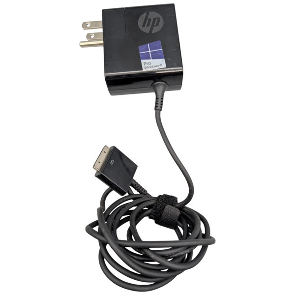 An overhead shot of a black, square HP AC adapter and its attached power cable against a white background. The adapter features a silver HP logo and a blue Windows 8 Pro sticker on its face, with a two-prong North American power plug extending from the top left side. A thin, black cable extends from the bottom of the adapter, leading to a coiled section secured by a black Velcro strap. The cable terminates in a wide, proprietary HP tablet connector.