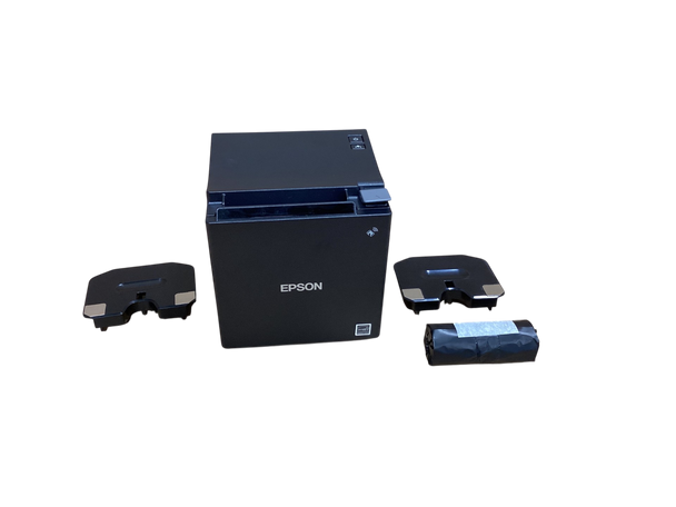 A matte black, cube-shaped Epson thermal receipt printer is centered on a plain, light brown cardboard surface. The front face features the "EPSON" logo in white text, a small wireless symbol near the top right, and an Energy Star sticker in the bottom right corner. On the top panel, two small buttons—a power button and a feed button—are visible on the right side next to a gray paper release lever. Flanking the printer are two identical, detached black plastic brackets or covers with silver metallic accents on their edges. A tightly wrapped, small roll of black thermal paper secured with white tape lies to the right of the printer.