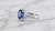 The ring displayed elegantly atop a minimalist white porcelain or micro-suede display element, angled slightly to highlight the raised profile of the setting and the intricate basket work beneath the sapphire.