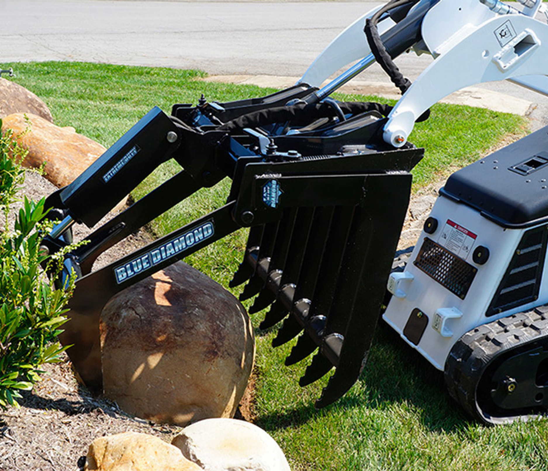 front right angled view of a mini skid steer on grass with a blue diamond mini skid steer root grapple attachment on the front grabbing a log