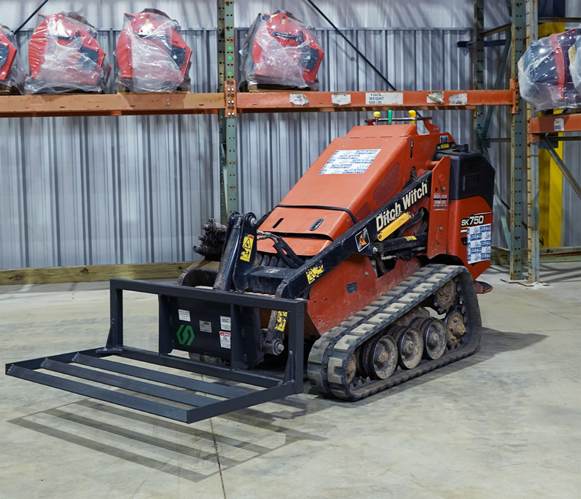 right angled view of a mini skid steer parked in a warehouse with a cid mini skid steer land leveler attachment on the front