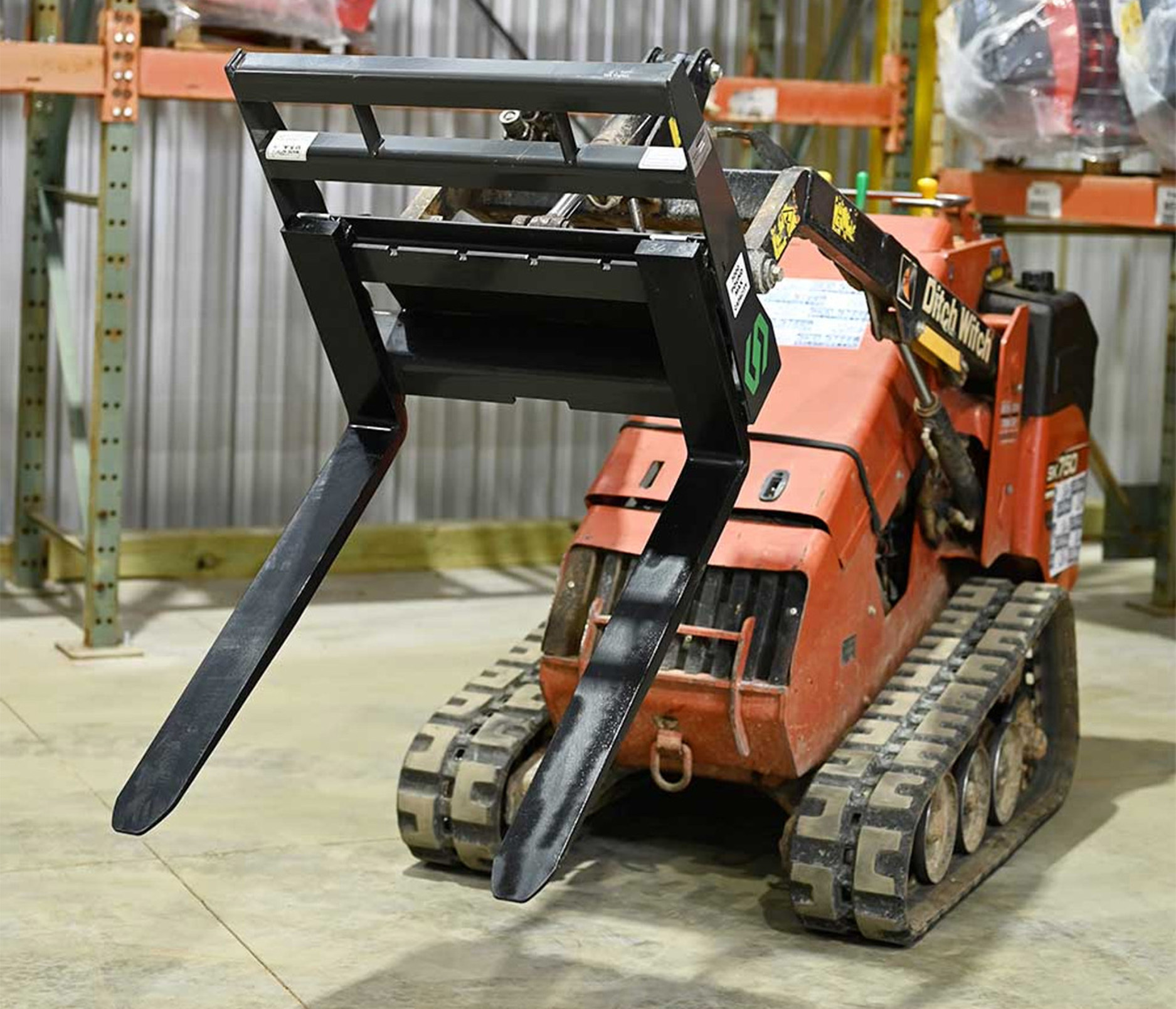 right angled view of a mini skid steer parked in a warehouse with a cid mini skid steer pallet fork attachment on the front