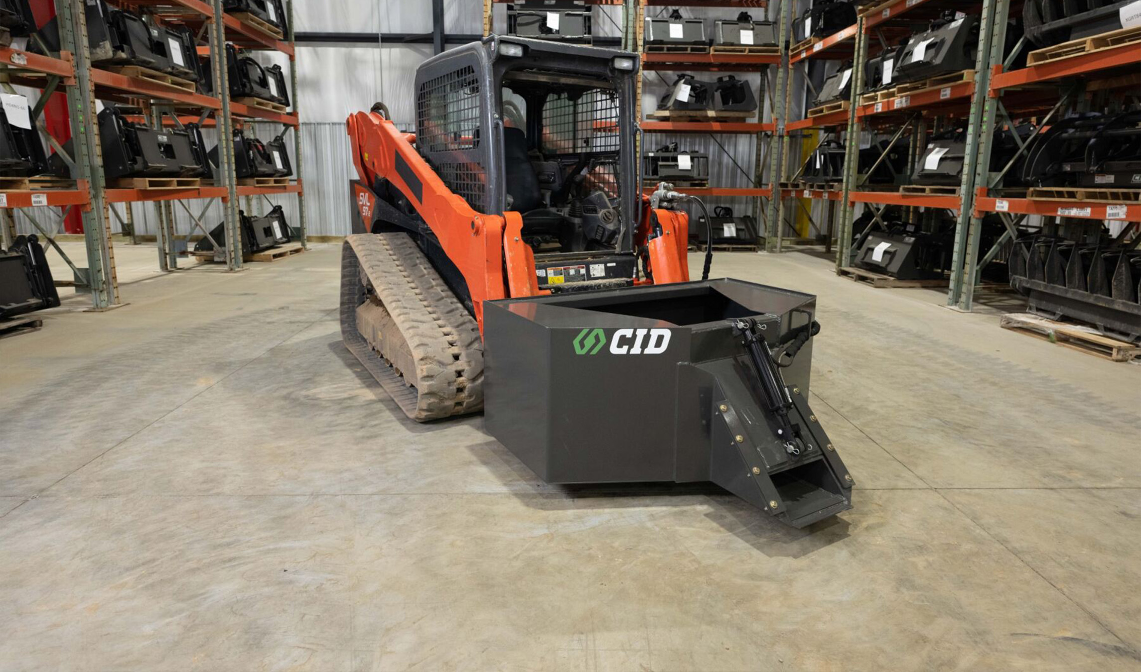front angled view of a skid steer parked in a warehouse with a CID Concrete dispensing bucket attachment on the front