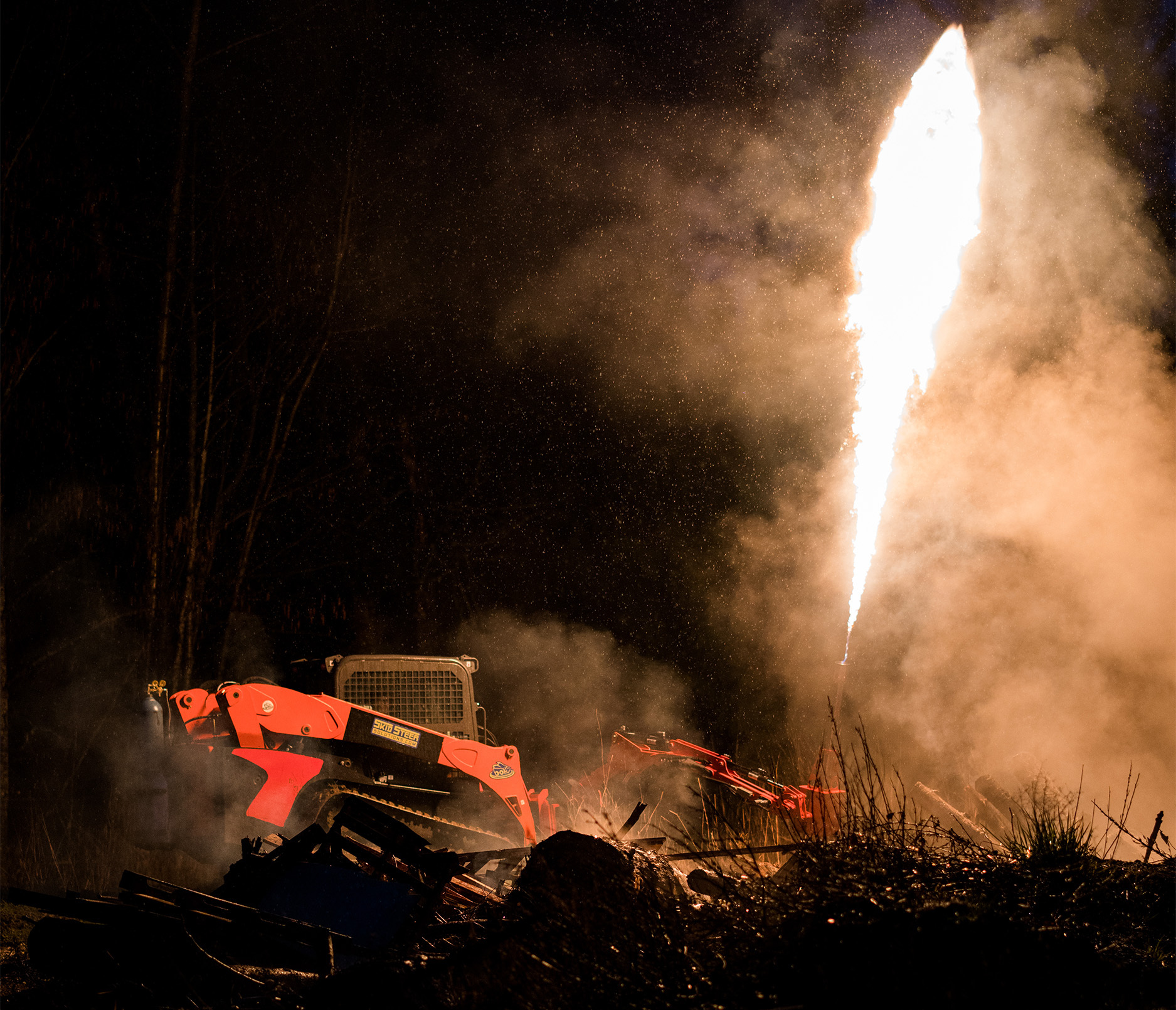 side view of a skid steer in a field at night time in a wooded area with an eterra flame thrower mounted on the front shooting fire