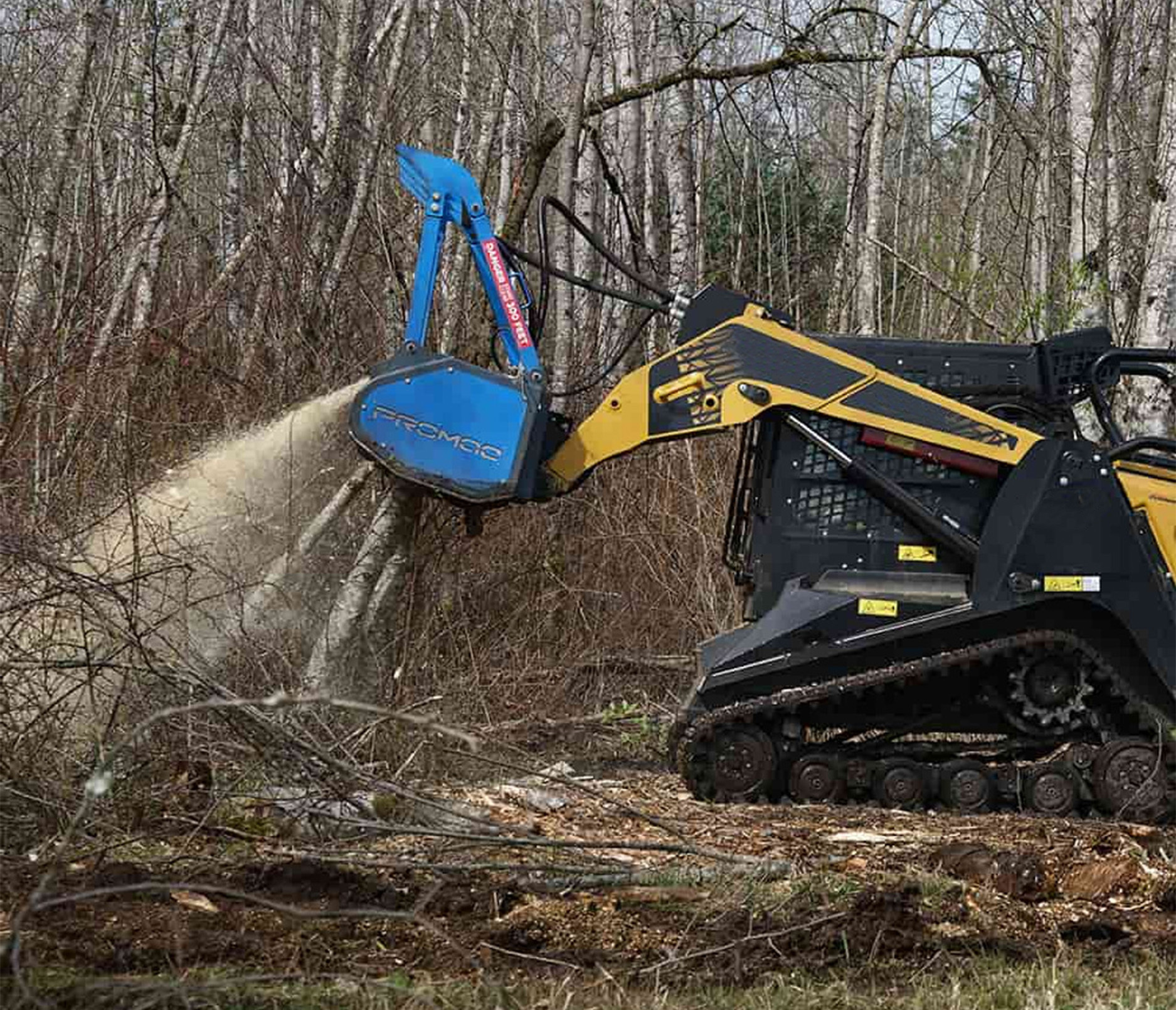 left side profile view of a skid steer in a forested area with a Promac HSM60 Attachment on the front, mulching down a tree